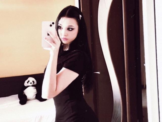 MaganSplonskowski with long black hair, wearing a black shirt, taking a mirror selfie, showing her ass, with a panda stuffed toy on the bed.
