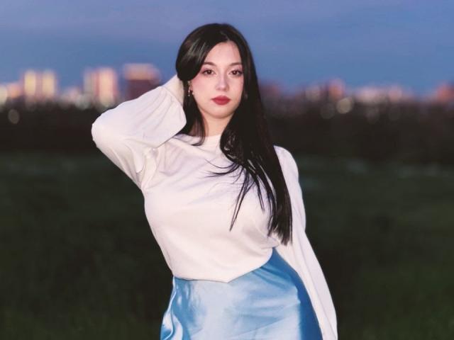 MaganSplonskowski with long black hair, white long-sleeve shirt, and blue satin skirt, standing with one hand in her hair, looking at the camera with red lipstick.