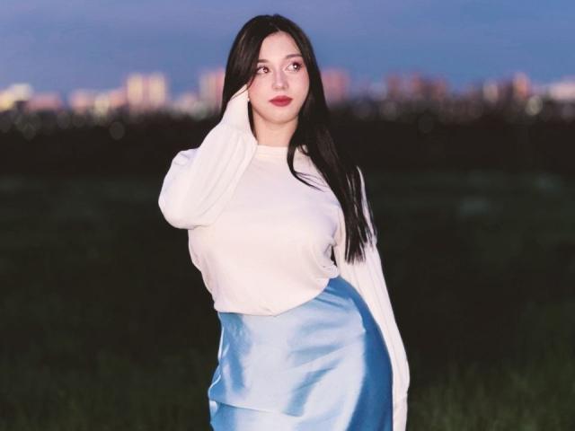 MaganSplonskowski with long black hair, white long-sleeve shirt, and light blue satin skirt, touching her cheek, standing in a grassy field at dusk, sky in blue.