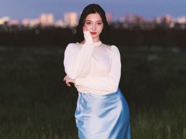 MaganSplonskowski with long dark hair, white long-sleeve shirt, and shiny blue satin skirt, hand on cheek, standing in grassy field, city lights in background, dusk.