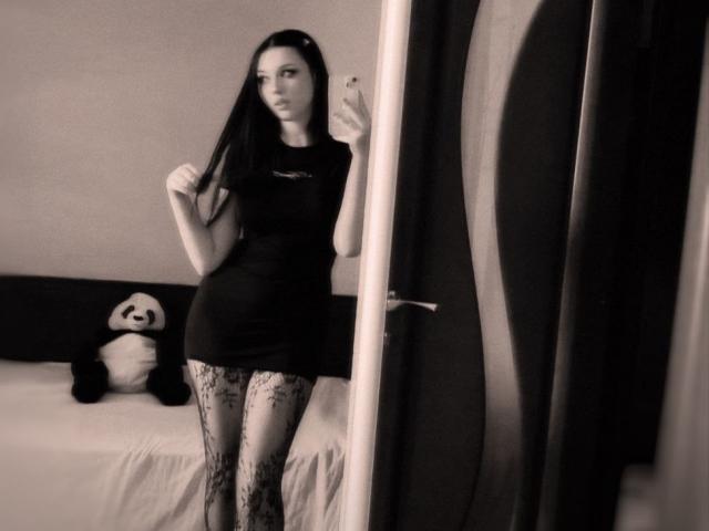 MaganSplonskowski with long dark hair, black dress, and lace-patterned tights, standing in front of mirror, holding phone, panda stuffed toy on bed to the left.