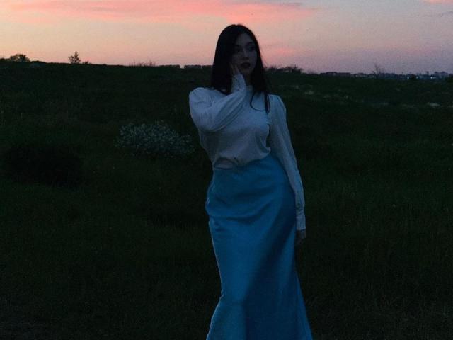 MaganSplonskowski has dark hair, a white long-sleeve top, and a blue skirt, standing in a dark grassy field at sunset, with pink and orange sky in background.