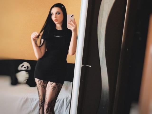 MaganSplonskowski stands in front of a mirror, holding her long black hair with one hand, wearing a tight black dress and lace stockings, showing her ass.