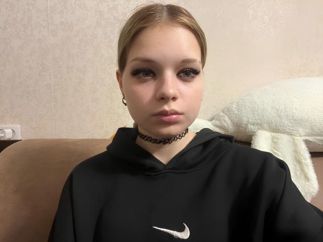 PingKipper met lichte huid, bruin haar en dikke wimpers, draagt een zwarte Nike hoodie en een zwarte choker, zittend op een bruine bank en kijkend naar de camera..