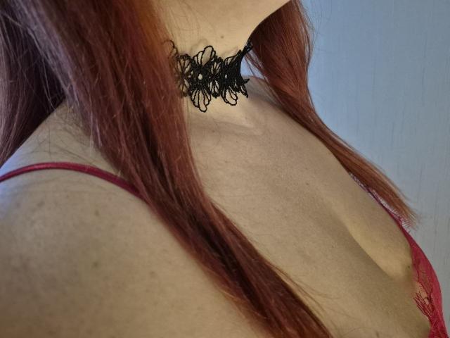 Dinastiia's red bra, showing her tits, with a black lace choker, and red hair falling down. Her pale skin is visible. Her shoulder is slightly turned.