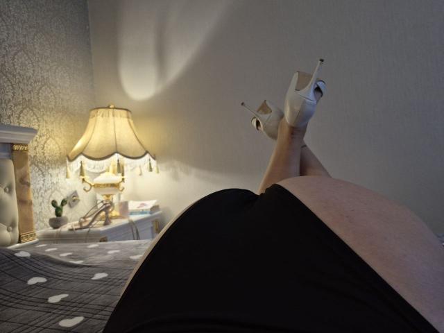 Dinastiia's legs crossed at the ankles, wearing white high heels, with a black skirt, her butt in the air, on a bed with a plaid blanket.