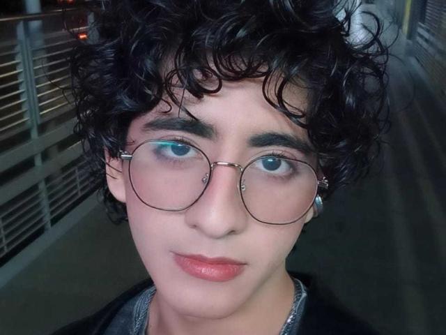 ElliotNoir with curly black hair, wearing round glasses, light pink lipstick, and a black jacket, showing a calm expression, standing in a dimly lit area.