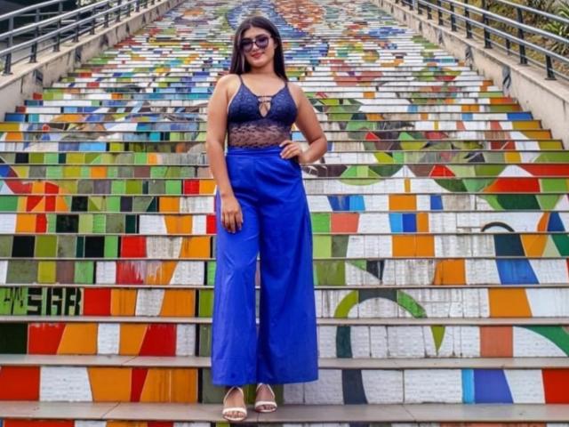 Daphne, cheveux foncés, portant un haut en dentelle violette, un pantalon bleu taille haute, debout sur des marches colorées, sandales marron, souriante, portant des lunettes de soleil, une main sur la hanche..
