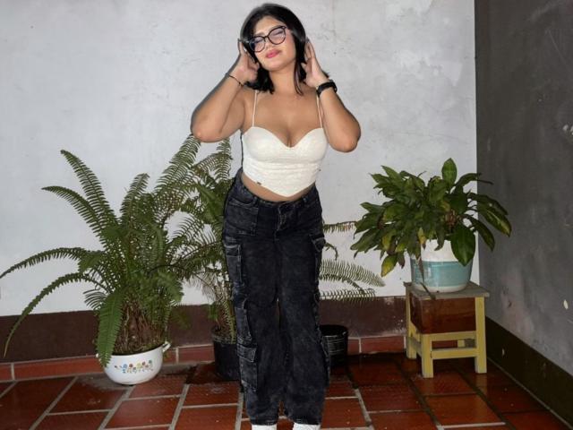 DaphneBrow a les cheveux noirs, porte des lunettes et un haut blanc sans bretelles qui dévoile sa poitrine. Elle porte un pantalon noir et se tient près de plantes vertes sur un sol carrelé..