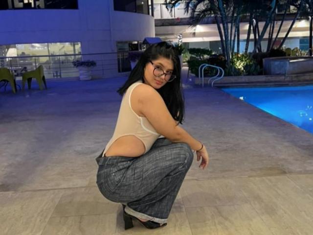 DaphneBrow sitzt am Beckenrand, trägt eine Brille, ein durchsichtiges weißes Oberteil und graue Hosen, wobei ihr Hintern und ein kleiner Po zu sehen sind, und trägt schwarze High Heels..