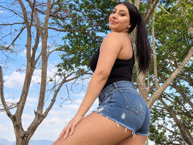 CatalinaRivero with dark ponytail, black tank top, and frayed denim shorts, showing her ass, standing outdoors with trees and blue sky in background. Her skin is tan.