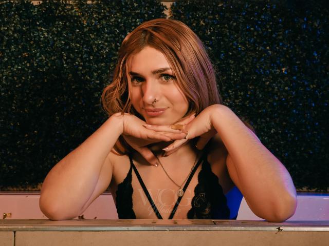 AndyDupont with shoulder-length brown hair, black lace top, hands under chin, subtle smile, blue eyes, small nose ring, necklace, leaning on wooden surface, soft lighting, cozy expression.