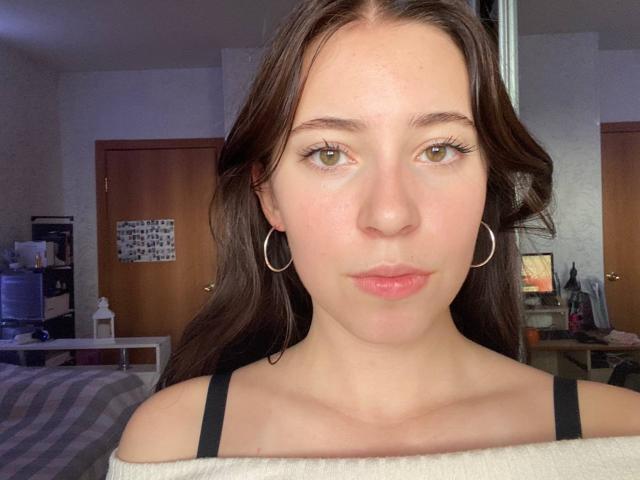 Alene Falcioni, avec des yeux verts, des cheveux bruns ondulés, des lèvres roses, de petites boucles d'oreilles, et des bretelles spaghetti noires, portant un haut blanc à une épaule, regardant directement l'appareil photo..