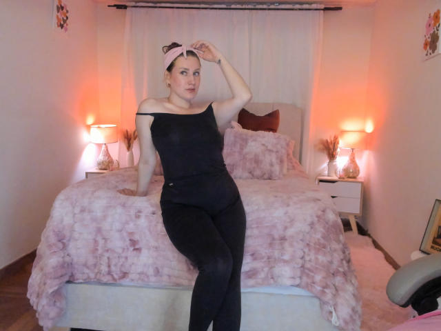 KatyaRogersX, with her tits barely visible through a black off-shoulder top, sits on a bed in a pink and black room, hand on head, wearing pink headband.