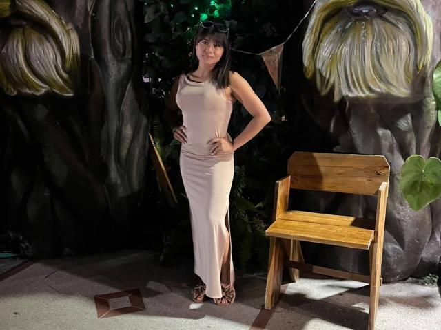 AshlyLee stands in a forest-themed setup, wearing a beige, sleeveless, form-fitting dress, showing her tits, with hands on hips, and black sandals, next to a wooden chair.