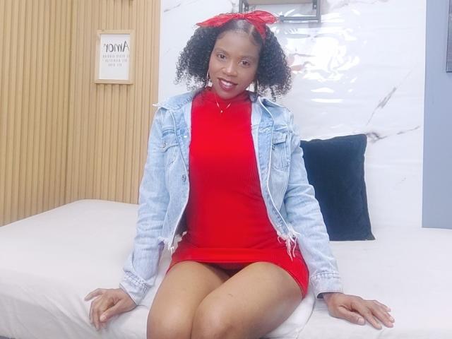 SarahiPusey, with curly black hair and red bow, wears a red dress, denim jacket, and sits on a bed, showing her red pussy through her dress, smiling.