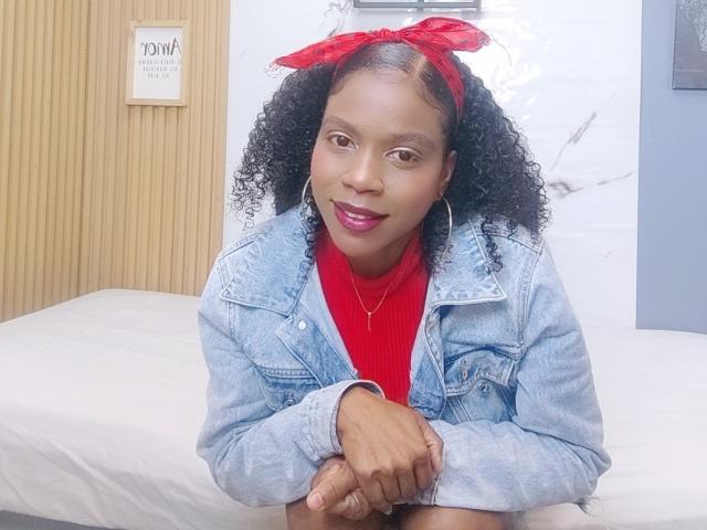 SarahiPusey with curly black hair, red bandana, red turtleneck, denim jacket, and red lipstick, sitting on a bed, smiling with hoop earrings and crossed arms.
