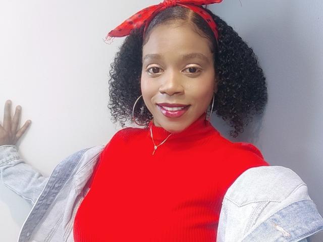 SarahiPusey with curly black hair, red bandana, red turtleneck, large hoop earrings, and light denim jacket, smiling with red lipstick, hand on white wall.
