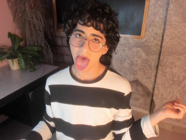 ElliotNoir with curly black hair, round glasses, and a tongue out, wearing a black and white striped shirt, looking at the camera, right arm raised.