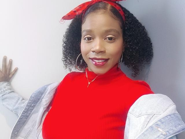 SarahiPusey with curly black hair, red headband, red turtleneck, and denim jacket, showing her tits, smiling with red lipstick, wearing hoop earrings, standing against a white wall.