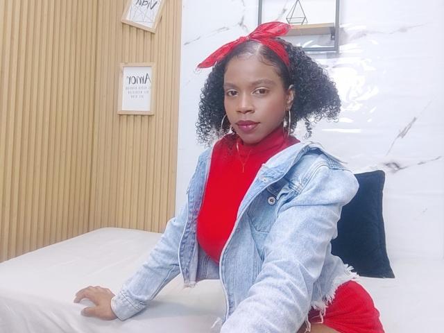 SarahiPusey with curly black hair, red bow, red turtleneck, denim jacket, and red pants, leaning on white bed, wearing hoop earrings, with a confident, sexy expression.