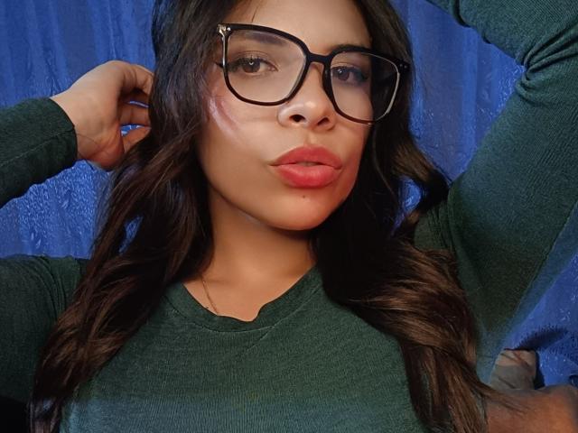 VioletaStone with glasses, pouty red lips, wavy brown hair, green sweater, hand in hair, blue background, slightly flushed cheeks, soft light.