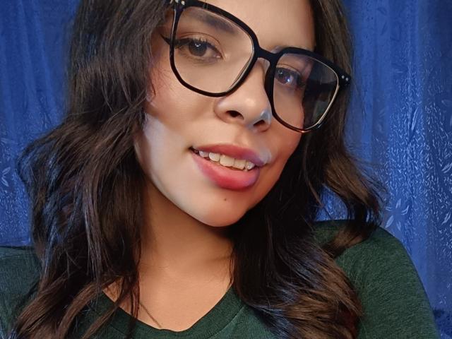 VioletaStone with wavy brown hair, wearing black glasses and a green shirt, has pink lipstick and a small moleskin on her right cheek, smiling.