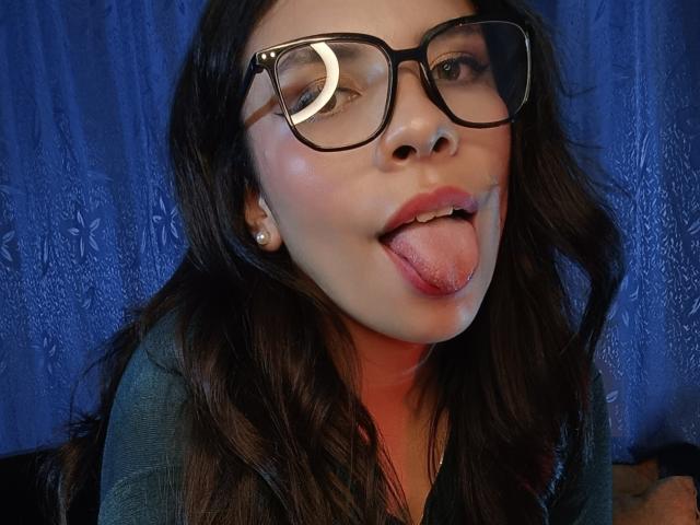 VioletaStone with dark wavy hair, black glasses, and pearl earrings, sticking out her tongue. She has a light skin tone and is wearing a green top.