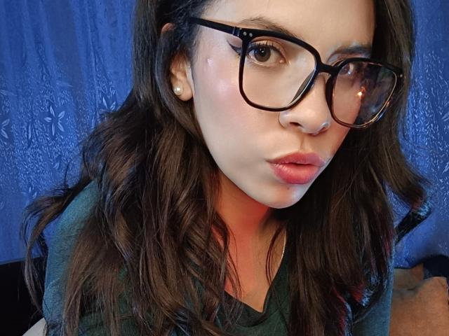 VioletaStone with puffy pink lips, wearing black glasses, green shirt, and pearl earrings, has wavy brown hair and a fair skin tone, pouting slightly.