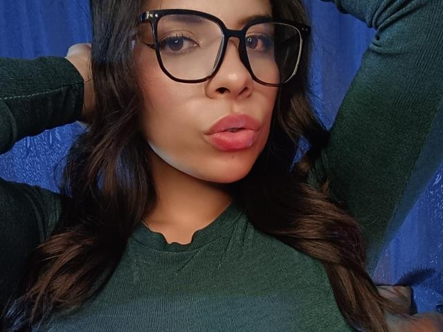 VioletaStone with dark wavy hair, wearing black glasses, green sweater, pouting pink lips, hands behind head, blue background, medium skin tone, attractive face.