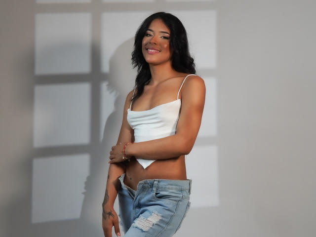 AgataDolly with dark hair, smiling, wearing a white crop top, showing her tits, and blue ripped jeans, standing against a white wall with shadow patterns.