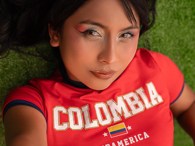 MelizQueen with brown eyes, red makeup on eyelids, lying on green grass, wearing red Colombia jersey, glossy brown lips, dark wavy hair, slight smile, cute, sexy.