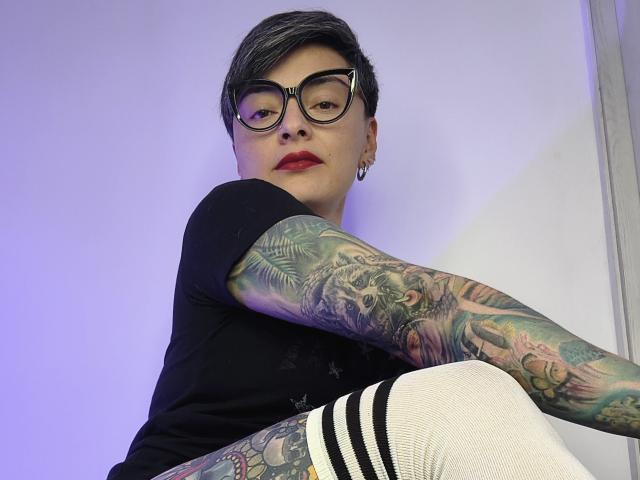 KatParadise, con i capelli corti grigi, occhiali, rossetto rosso, una maglietta nera e calze bianche con strisce nere, mostra braccia tatuate colorate, guarda in basso, in una posa rilassata..
