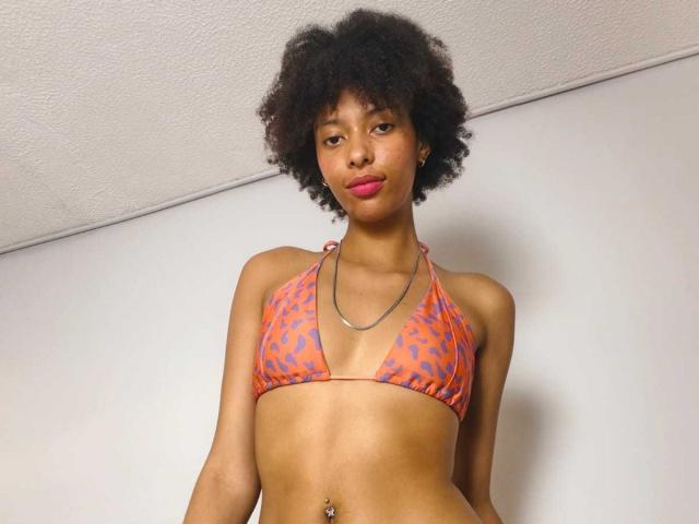 KiaraKamau with a small orange and blue leopard print bikini, showing her boobs and a belly button piercing, has dark curly hair and pink lipstick, looking down.