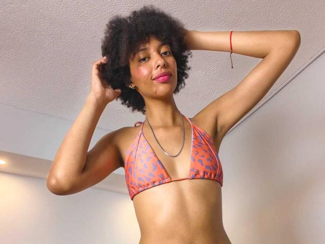 KiaraKamau with curly black hair, pink lipstick, wearing an orange leopard-print bikini top, silver necklace, and red bracelet, posing with hands in her hair.