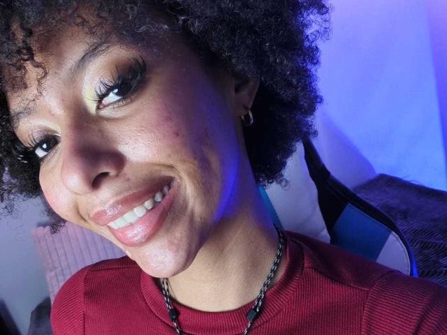 KiaraKamau with curly black hair, smiling, wearing a red sweater, chain necklace, and hoop earring, has glossy pink lips and long eyelashes, in a well-lit room.