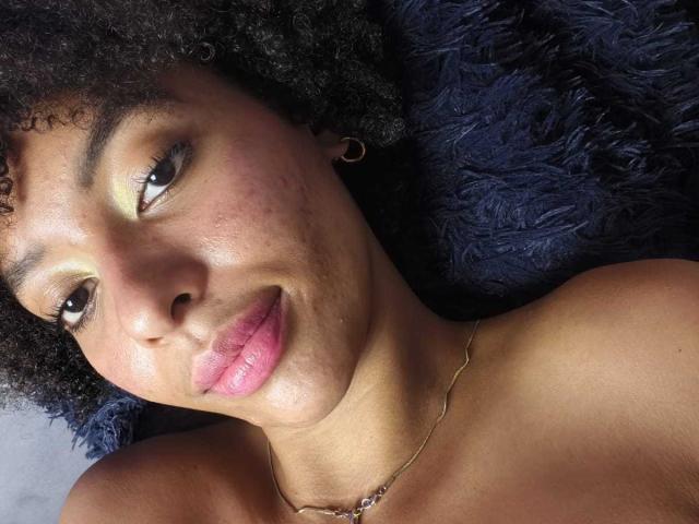 KiaraKamau's face with curly black hair, brown eyes, yellow eyeshadow, pink lipstick, slight smile, small gold hoop earrings, and a thin gold necklace, lying on a dark blue blanket.