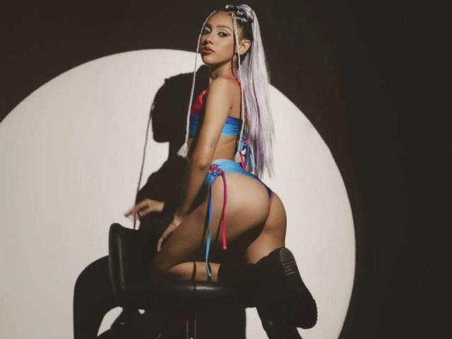GabySmiith met lange witte vlechten, die een blauwe en rode string bikini draagt, knielend op een kruk, waarbij haar ronde billen te zien zijn, en ze kijkt over haar schouder, in een donkere kamer..