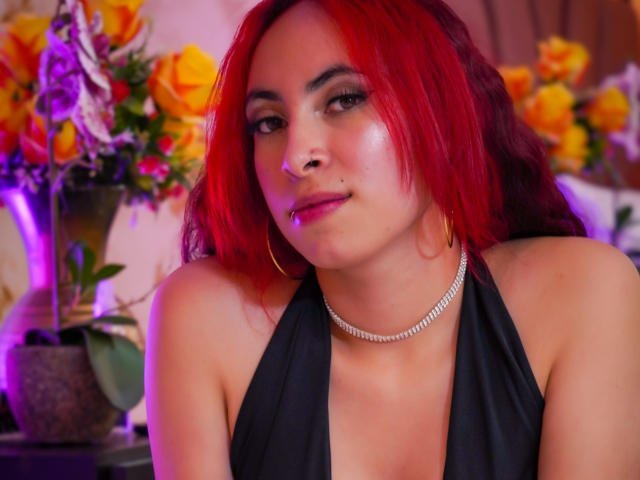 Anya Carter, con i capelli rossi, un top nero a fascia, una collana di diamanti e orecchini a cerchio, mostra un sorriso appena accennato, con un piccolo piercing al naso e rossetto rosa, seduta di fronte a dei fiori..
