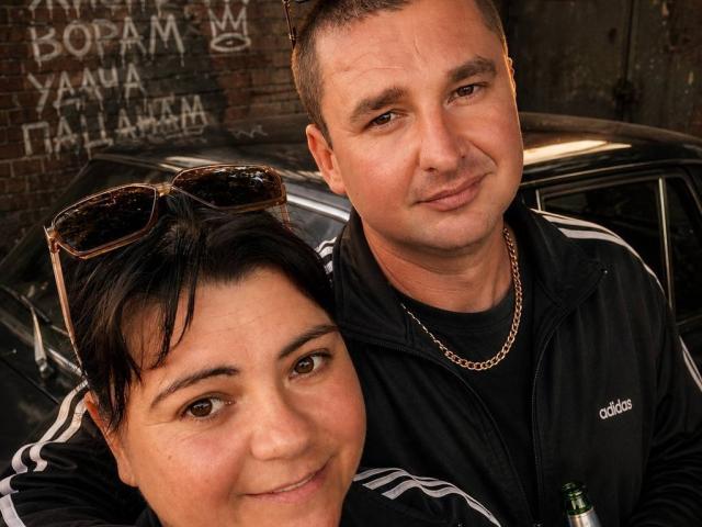 Mariyna69 with short black hair and brown eyes, wearing a black Adidas jacket, stands next to a man with a shaved head and chain, both smiling. Car in background.