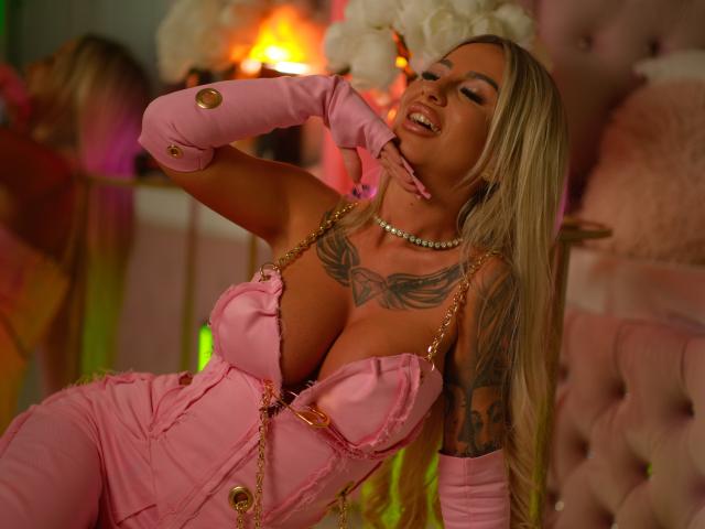 CelineDian with blonde hair, pink dress, gold buttons, and gloves, has large tits, tattoos, and a pearl necklace, eyes closed, hand near mouth, sitting on pink tufted chair.