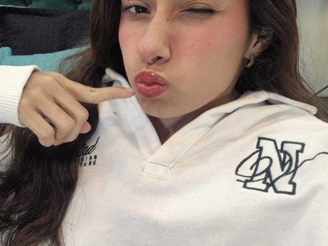 MiliBlom with puckered lips, pointing at them with her finger, wearing a white hoodie with a black "AV" logo, her face slightly flushed, brown hair, and subtle makeup.