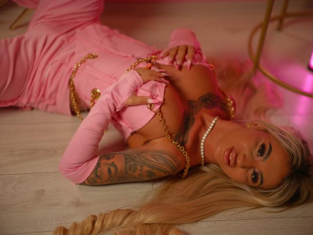 CelineDian is lying on a wooden floor, wearing a pink dress with gold chains, exposing her big tits, and has long blonde hair, tattoos on her arm, and a pearl necklace.