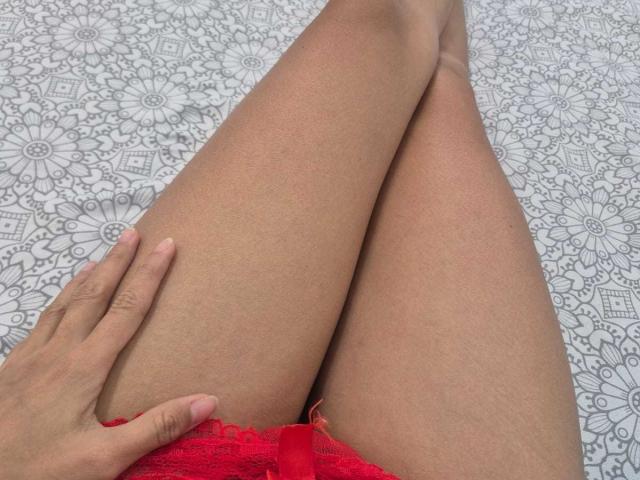 MiliBlom's red lace panties are visible, with her hand resting on her upper thigh. Her tan legs are crossed, showing smooth skin. A patterned white bedsheet is underneath.