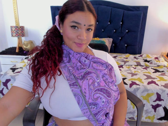 DarsshaHara with big tits, wearing a white top and purple paisley scarf, curly red hair, pink earrings, sitting in a chair, smiling, bedroom with blue headboard and patterned bed.