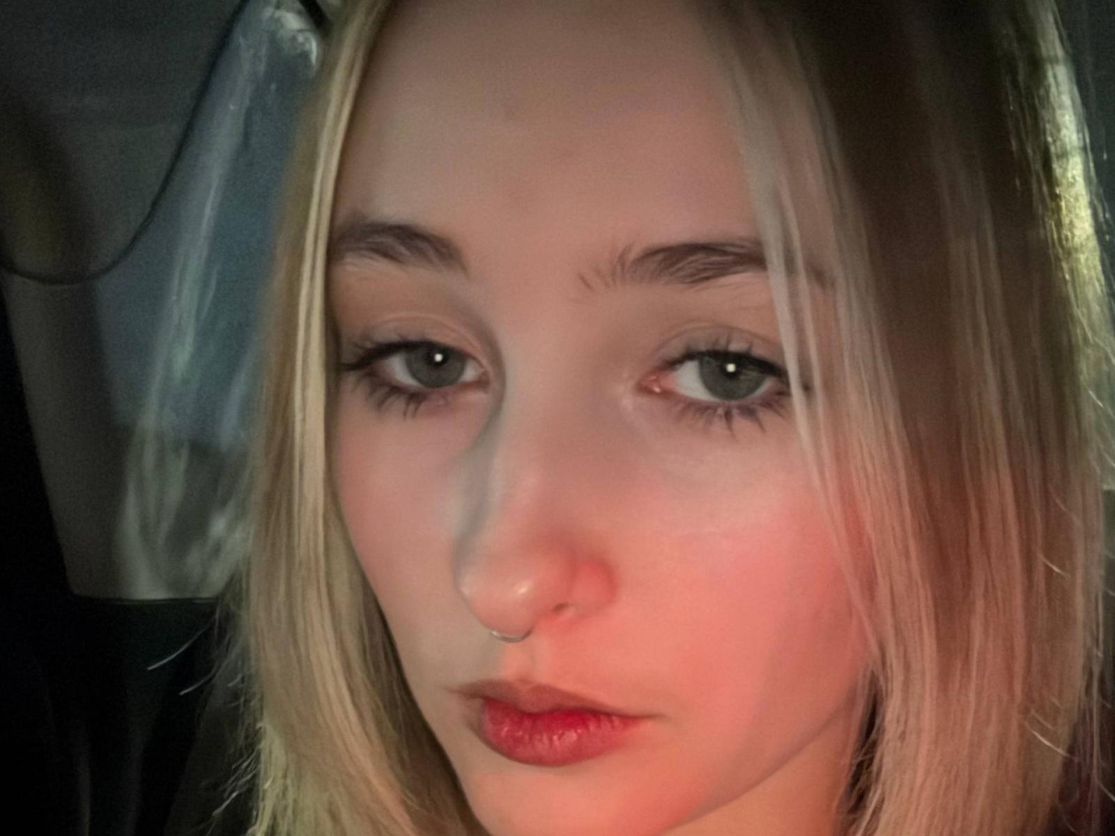 MarishaSchink's face with blonde hair, green eyes, nose ring, and red lipstick, looking serious in a dimly lit setting, with soft light highlighting her right cheek.