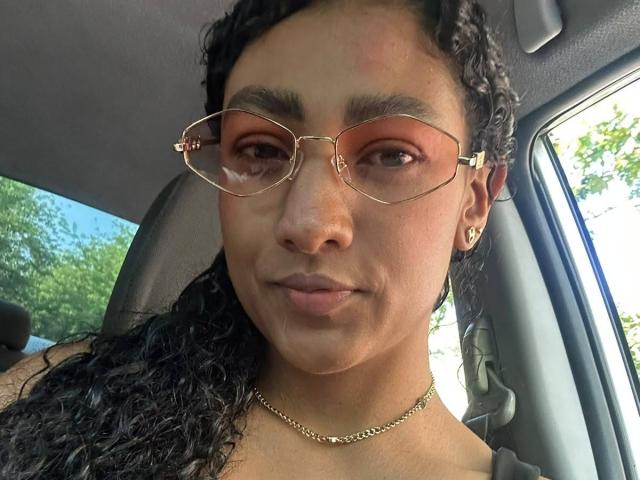 NatalyBlonder has curly black hair, tan skin, and wears gold glasses with brown lenses, a gold chain necklace, and a diamond earring. She's sitting in a car.