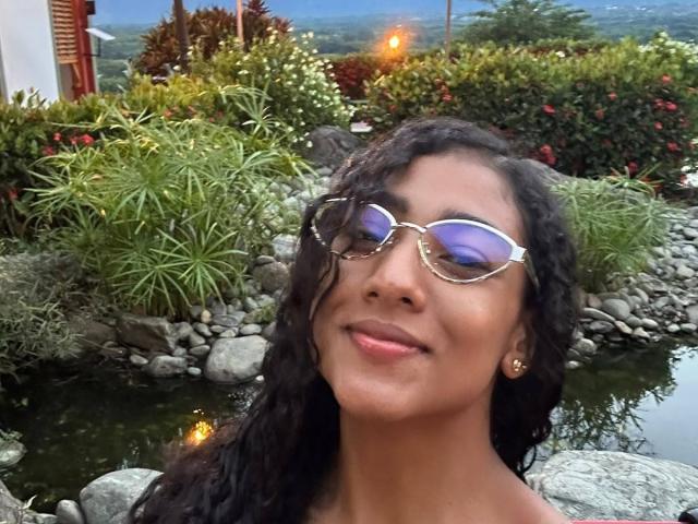 NatalyBlonder with curly black hair, wearing reflective sunglasses and gold earrings, smiling at the camera, standing by a rocky water feature. Her brown skin is glowing.