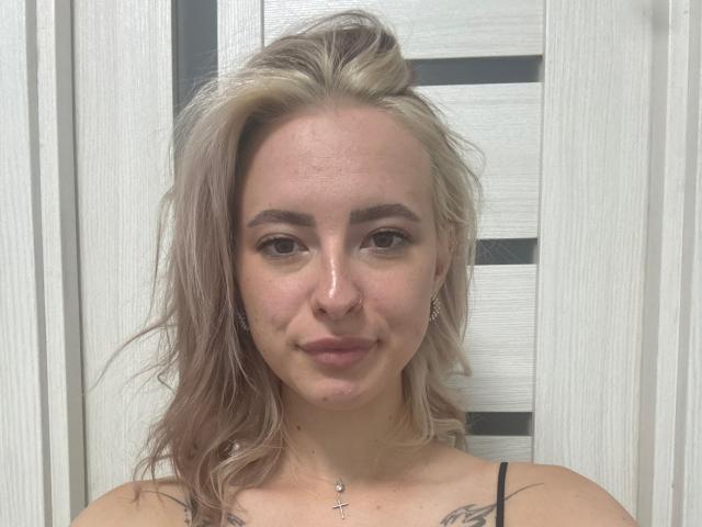 MarlenaPort tem cabelo loiro e ondulado, pele clara, nariz pequeno, sobrancelhas escuras, tatuagens discretas, um colar com uma cruz e um sorriso leve. Ela está vestindo uma blusa preta com alças finas..