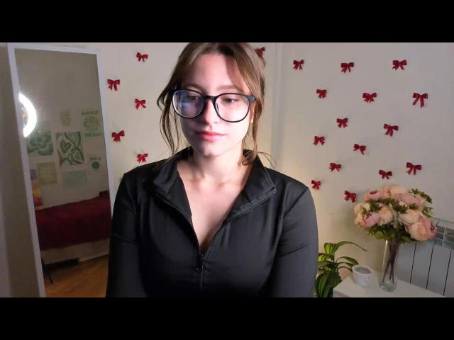 LupeYing, with glasses and brown hair, wearing a black zip-up shirt, has small tits and a cute face, standing in a room with red bows on the wall.